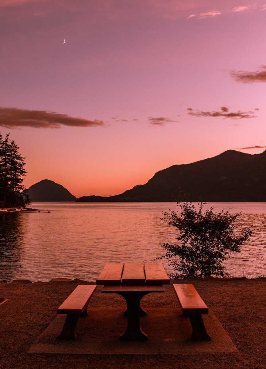 brown wooden table and bench near body of water during sunset